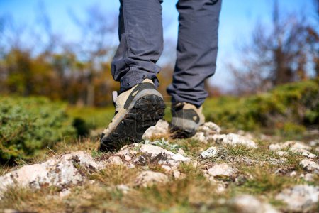 Téléchargez les photos : Legs of a hiker in trekking boots walking in mountains closeup shot. Feet of walking tourist wearing trekking shoes on a rocky road captured from behind. Hiking male wearing pants and boots walk - en image libre de droit