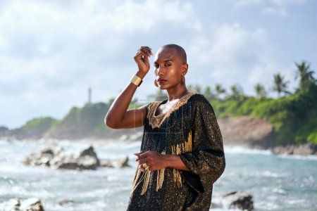 Photo for Androgenous black fashion model stands on scenic tropical beach portrait. Non-binary ethnic person wearing makeup, luxurious jewellery poses with hands at exotic seaside location. Trans sexual, pride - Royalty Free Image