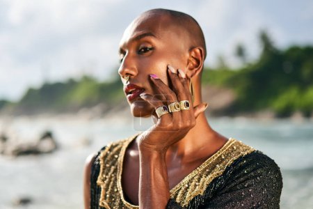 Photo for Outrageous gay black man in luxury gown, jewelry poses on scenic ocean beach. Gender fluid ethnic fashion model in a posh dress, accessories looks at camera touches face with hand and rings on fingers - Royalty Free Image