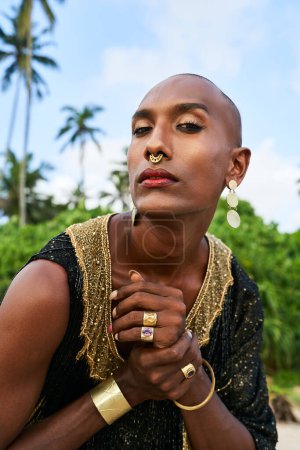Photo for Flamboyant gay black man with luxury golden jewelry poses on scenic ocean beach. Gender fluid ethnic fashion model brass rings, bracelet, earrings, accessories looks stands. Sophisticated posture. - Royalty Free Image