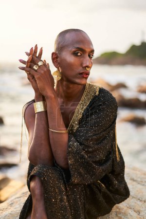 Non-binary black person in luxury dress sits on rocks in ocean. Trans ethnic fashion model wearing jewelry in a posh gown, poses in tropical seaside location. Divine feminine human. Diversity concept.