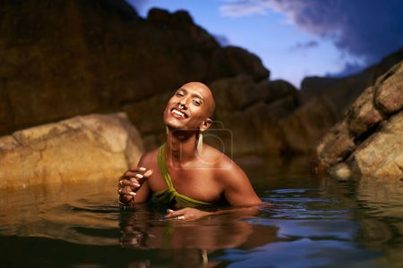 Photo for Smiling gender fluid black person poses in natural still water pool. Queer ethnic fashion model in open dress, brass jewelry with gems standing gracefully in the middle of a rocky lake at night. - Royalty Free Image