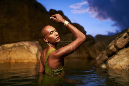 Photo for Non-binary person of color poses in water, shows jewelry inside scenic creek at night. Lgbtq ethnic graceful fashion model wear brass rings with gems, nose-ring, earrings, bracelets immersed in pond. - Royalty Free Image