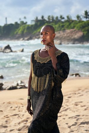 Photo for Trans sexual ethnic fashion model in long posh dress and accessories in elegant posture looks at camera. Epatage gay black man in luxury gown poses, touches face on scenic an ocean beach. - Royalty Free Image
