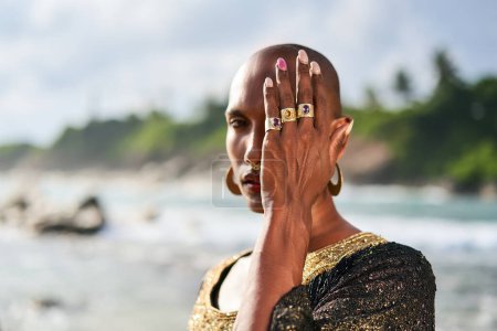 Photo for Outrageous gay black man in luxury gown, jewelry poses on scenic ocean beach. Gender fluid ethnic fashion model in a posh dress, accessories looks at camera covers face with hand and rings on fingers. - Royalty Free Image