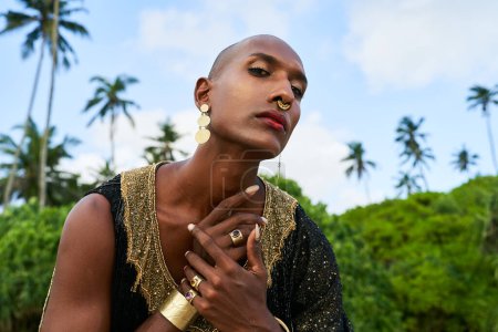 Photo for Gorgeous gay black man in luxury gown, jewelry poses on scenic ocean beach . Non-binary ethnic fashion model wears long posh dress golden rings, earrings, bracelets, looks at a camera. - Royalty Free Image