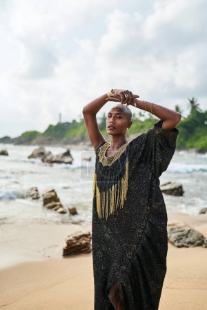 Photo for Epatage lgbtq black male posing with hands up on camera on scenic ocean beach. Non-binary ethnic fashion model in long posh dress wears jewellery stands gracefully on sea shore and a lighthouse. - Royalty Free Image
