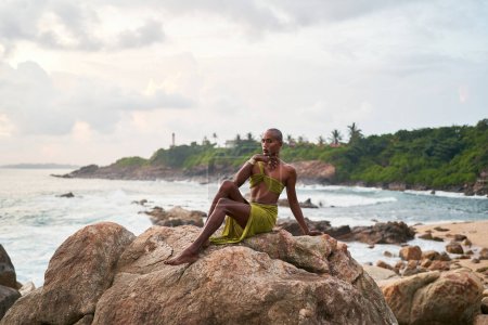 Photo for Non-binary black person poses gracefully sitting on top of rock in ocean at sunset. Trans ethnic divine fashion model poses in open tropical dress on peak by a storm at dusk against sunrise. LGBT - Royalty Free Image