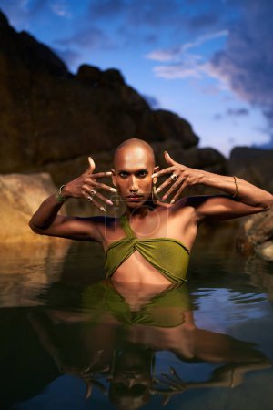 Photo for Androgynous bipoc lgbtq model poses in water inside natural pool at night. Non-binary person shows jewelry - rings with gems on fingers, brass nose ring, golden earrings, bracelets, stands in a pond. - Royalty Free Image