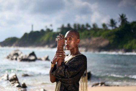 Photo for Flamboyant gay black man in a luxury gown poses on scenic ocean beach. Gender fluid ethnic fashion model in long posh dress and accessories look at camera stands with sophisticated posture. - Royalty Free Image