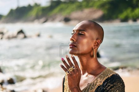 Photo for Trans sexual ethnic fashion model in posh dress and jewelry accessories in elegant posture touches neck. Epatage gay black man with luxury rings, earrings touches a face on scenic ocean beach. Pride. - Royalty Free Image
