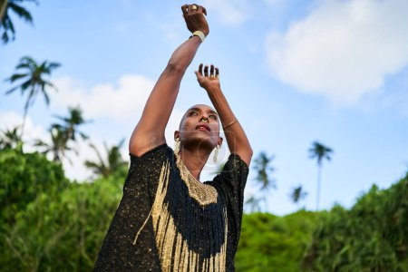 Photo for Epatage lgbtq black male posing with hands up on camera on scenic palm tree location. Non-binary ethnic fashion model in long posh dress wears jewellery stands gracefully on a tropical spot. - Royalty Free Image