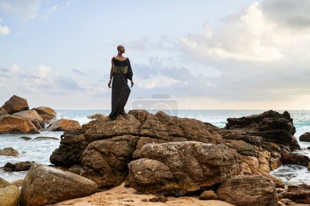 Photo for Androgynous black fashion model poses on a scenic ocean beach . Non-binary ethnic fashion model in long posh dress and accessories stands on rocks by sea against moody sky in dull cloudy weather - Royalty Free Image
