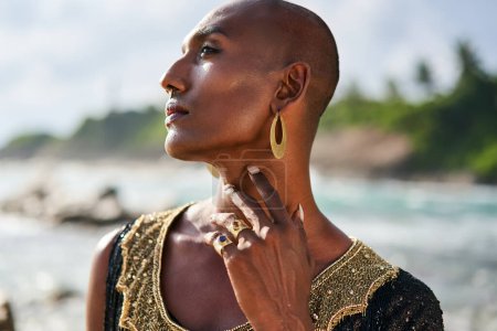 Photo for Trans sexual ethnic fashion model in long posh dress and accessories in elegant posture touches neck. Epatage gay black man in jewelry poses, touches neck on scenic an ocean beach. Pride, close up - Royalty Free Image