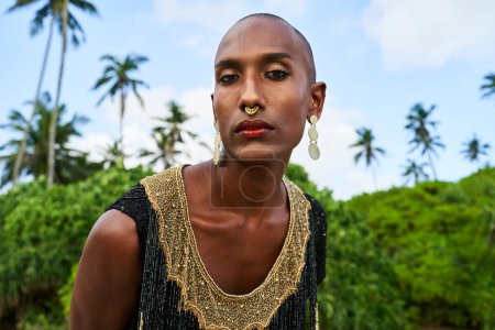 Photo for Gorgeous gay black man in luxury gown, brass jewelry poses on scenic ocean beach. Non-binary ethnic fashion model with golden rings, earrings, bracelets accessories looks at a camera. Pride month. - Royalty Free Image