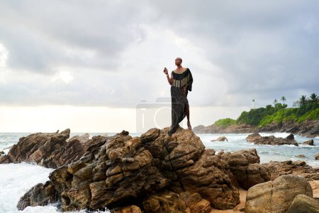 Photo for Trans sexual black person poses on scenic ocean beach like goddess. Lgbtq ethnic fashion model in long posh dress and accessories stands on rocks by sea against moody sky in dull cloudy weather - Royalty Free Image