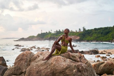Photo for Gender fluid black person poses gracefully sitting on high rock above ocean at sunset. Androgynous ethnic fashion model in dress on rocky hill by a storm at dusk. Pride month diversity concept. - Royalty Free Image