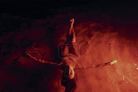Foto de La figura femenina serena descansa en una piscina iluminada por el rojo, la paz nocturna que resalta su delicada belleza. Su forma se desliza elegantemente, rodeada de resplandor escarlata de agua, creando una atmósfera mística. - Imagen libre de derechos