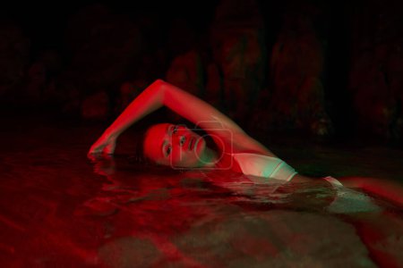 Téléchargez les photos : Modèle féminin nageant dans le lagon de nuit, éclairage rouge. Beauté et féminité se mêlent au cadre naturel de la piscine. Séance photo aquatique créative. Maillot de bain mouillé sur femme. Pose dans l'eau au crépuscule. - en image libre de droit