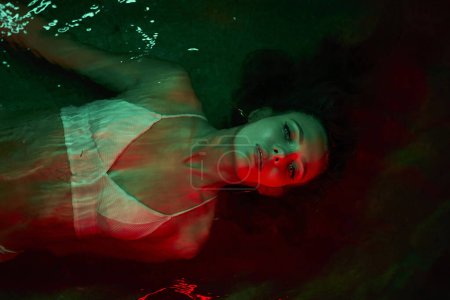 Photo for Female submerged in water under red light at night, creates surreal scene reflecting on mental state, split personality theme, and solitary contemplation for conceptual imagery. - Royalty Free Image