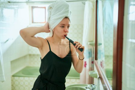 Téléchargez les photos : Femme s'engage dans les soins personnels, brosser les dents avec une brosse à dents motorisée dans le cadre de la maison, illustre l'importance de l'entretien de la santé buccodentaire. - en image libre de droit