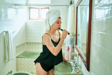 Téléchargez les photos : Reflet de dame coûteuse effectuant la routine de soins buccodentaires du matin, soies sur la brosse à dents, en face de l'évier de salle de bains et miroir avec serviette blanche enroulée autour de la tête. - en image libre de droit
