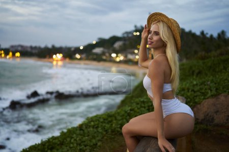 Photo for Young lady in swimwear relaxes on exotic coastline during sunset, luxury travel destination. Chic tourist contemplates serene seascape, tranquil evening by the sea, holiday relaxation. - Royalty Free Image