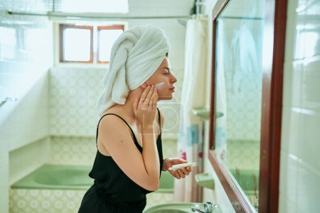 Photo for Towel-wrapped hair, retro tiles, skincare routine, mirror reflection capture the essence of daily beauty regimen. Woman in a vintage bathroom applies sunscreen to her face for skin protection. - Royalty Free Image