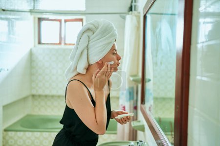 Photo for Skin care routine, mirror reflection at home spa. Woman in retro bathroom applies face cream, wrapped in towel. Female enjoys organic skincare product, self-care beauty regimen in vintage interior. - Royalty Free Image