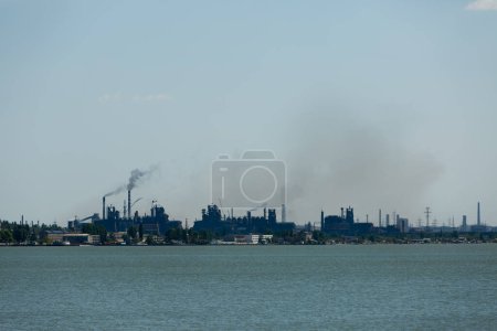 Photo for Industrial skyline dominated by metallurgical plant emissions, smoke billowing against sky, symbolizing eco pollution issues with water foreground. - Royalty Free Image