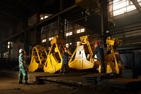Heavy equipment manufacturing process, teamwork with safety helmets. Industrial workers inspect yellow grapples in factory setting. Quality control in metal fabrication, engineered machinery parts.