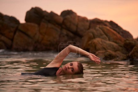 Female enjoys peaceful ocean swim near rocks. Woman in black swimwear relaxes in sea water at sunset. Serene beach bathing, wellness lifestyle. Harmony with nature, leisure activity, relaxation time.