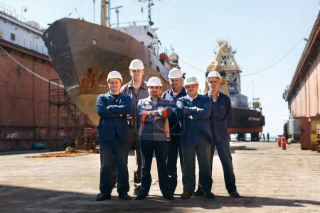 Marine engineers in helmets, repair crew with vessel. Team of shipyard workers stand proudly at dry dock. Dockyard laborers by ship hull, maritime industry, maintenance on sunshine day.