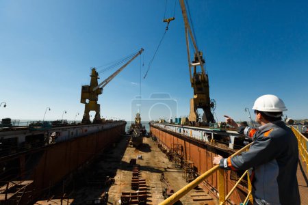 Photo for Maritime worker points, directs crane at shipyard under sunny sky. Engineer oversees vessel maintenance in dry dock. Industrial worker inspects, guides ship overhaul. - Royalty Free Image