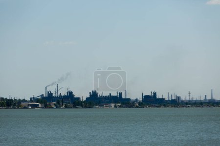 El humo se levanta contra el cielo azul, señalando la contaminación. skyline industrial a través del agua con la emisión de pilas en la planta metalúrgica. Complejo de fabricación emite subproducto, simbolizando cuestiones ecológicas.