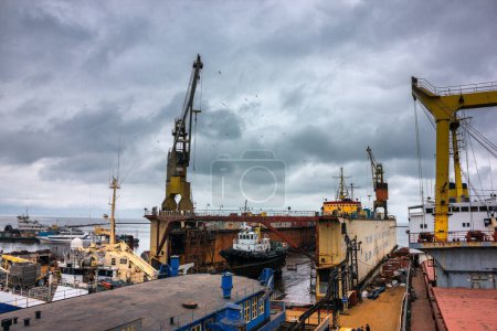 Tugboats assist in marine operations, heavy cranes lift equipment. Industrial ship repair yard with floating dock, vessels undergo maintenance. Overcast sky over maritime workplace, shipping industry