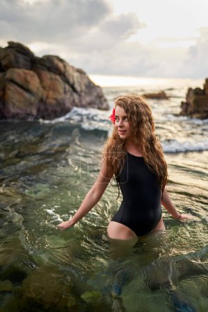 Alleinreisende erleben friedliches Baden im Meer. Elegante Frau mit roter Blume im Haar genießt das ruhige Bad im Meer bei Sonnenuntergang. Exotisches Reiseziel, goldene Stunde, Freizeitschwimmen, weibliche Schönheit am Meer.