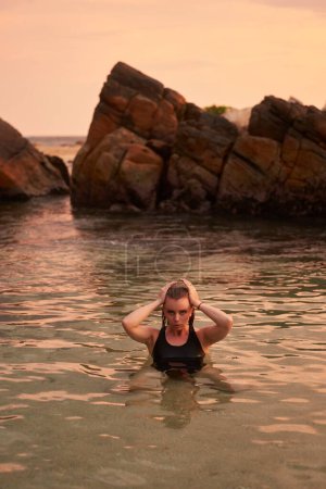 Bienestar, atención plena en un entorno natural. La mujer disfruta de un baño sereno al atardecer. Bañista femenina en el océano, tranquilidad. Abrazo de agua refrescante, rutina de fitness. Ocio, estilo de vida costero representado.
