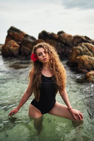 Sitzt am Meer, Felsenkulisse, Sonnenuntergang. Elegante Frau posiert im schwarzen Badeanzug, rote Blume schmückt das Haar. Ruhiges Porträt am Meer, Urlaubsreiselust. Bademoden-Modell in natürlicher Meeresumgebung.