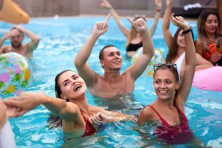 Amigos tienen fiesta en la piscina nocturna en una piscina villa privada. Jóvenes felices en traje de baño salpicando agua, bailando con flotadores y colchón inflable, pelota en resort de lujo.
