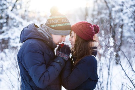 Young lovely couple warming up each others hands in snowy winter forest park.