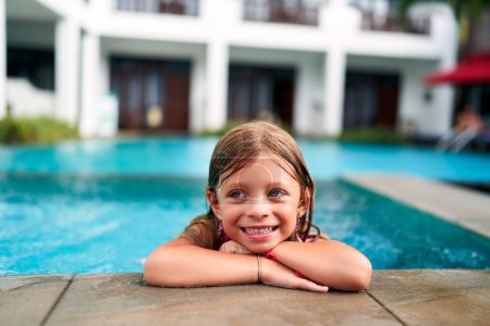 Das Kind genießt das sommerliche Wasservergnügen, schwimmt im kinderfreundlichen Pool. Lächelndes kleines Mädchen ruht am Beckenrand im sonnigen Resort. Urlaub, glückliche Kindheit, Familienreisen im Tropenhotel. Wasserspiel.