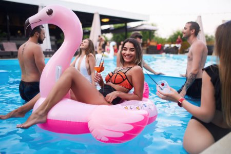 Mujer bonita en bikini traje de baño colgando en el colchón inflable de flamenco rosa en la fiesta de la piscina de la noche. Amigos de fiesta con cócteles en la piscina villa de vacaciones. Brindis de chicas, alegría con bebidas.