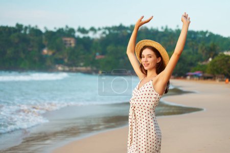 Smiling female enjoys tropical sun, waves, seaside vacation. Happy woman in straw hat, polka dot dress raises arms on sandy beach. Solo traveler explores coastal landscape, freedom concept.
