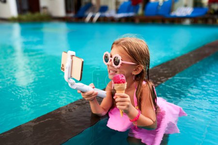 Child influencer films herself with smartphone, cheerful in summer resort setting. Little girl in pink swimsuit streams poolside, enjoys strawberry ice cream. Kids lifestyle, playful vlogger moment.
