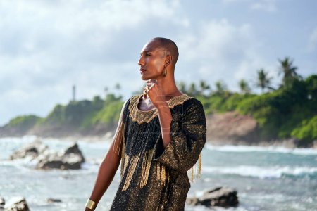 Empörende schwarze lgbtq Person im Luxuskleid posiert am malerischen Strand des Ozeans. Nicht-binäre ethnische Mode Modell in schickem Outfit steht anmutig an malerischer Lage in der Nähe des Meeres, Leuchtturm. Ein stolzer Monat.