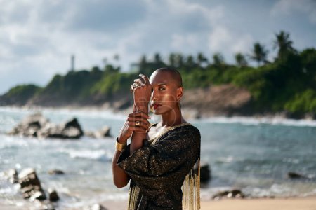 Flamboyant queer black male in luxury gown poses on scenic ocean beach. Trans sexual ethnic fashion model in long posh dress and accessories looks at camera, touches hands with rings and bracelet.