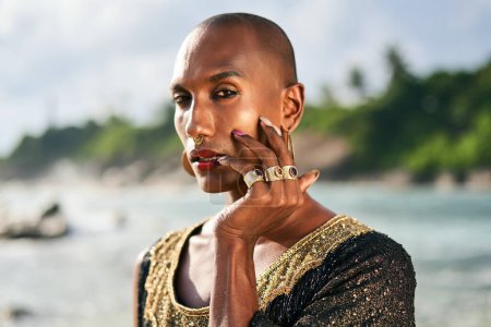 Photo for Outrageous gay black man in luxury gown, jewelry poses on scenic ocean beach. Gender fluid ethnic fashion model in a posh dress, accessories looks at camera touches face with hand and rings on fingers - Royalty Free Image