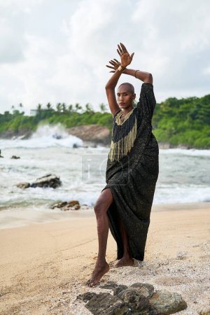 Epatage lgbtq schwarzer Mann posiert mit erhobenen Händen vor der Kamera am malerischen Strand des Ozeans. Nicht-binäre ethnische Mode-Modell in langen noblen Kleid trägt Schmuck steht anmutig an der Küste und einem Leuchtturm.