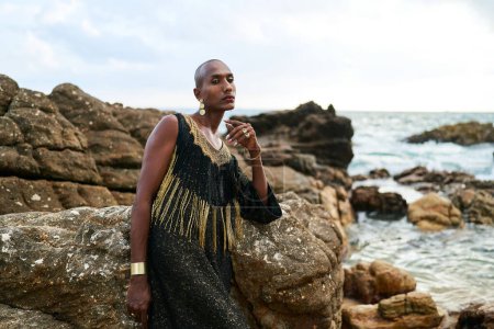 Queer black person in a luxury dress sits on rocks in ocean. Lgbtq ethnic fashion model wearing jewellery dressed in posh gown poses gracefully in tropical seaside location portrait. Pride month